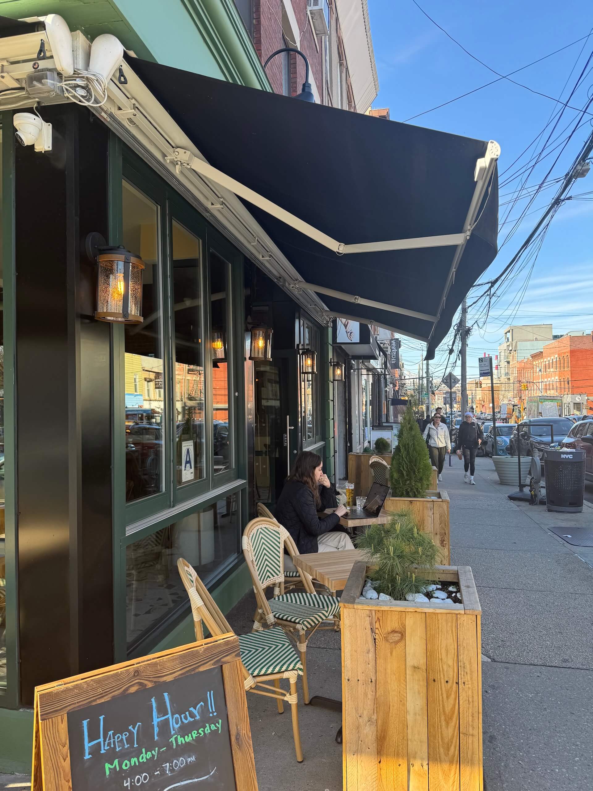 The green storefront under a black awning, with a Happy Hour sign and sidewalk tables